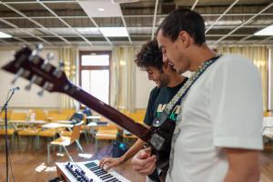 Two male students smiling and holding their guitars, while one is playing the piano as well
