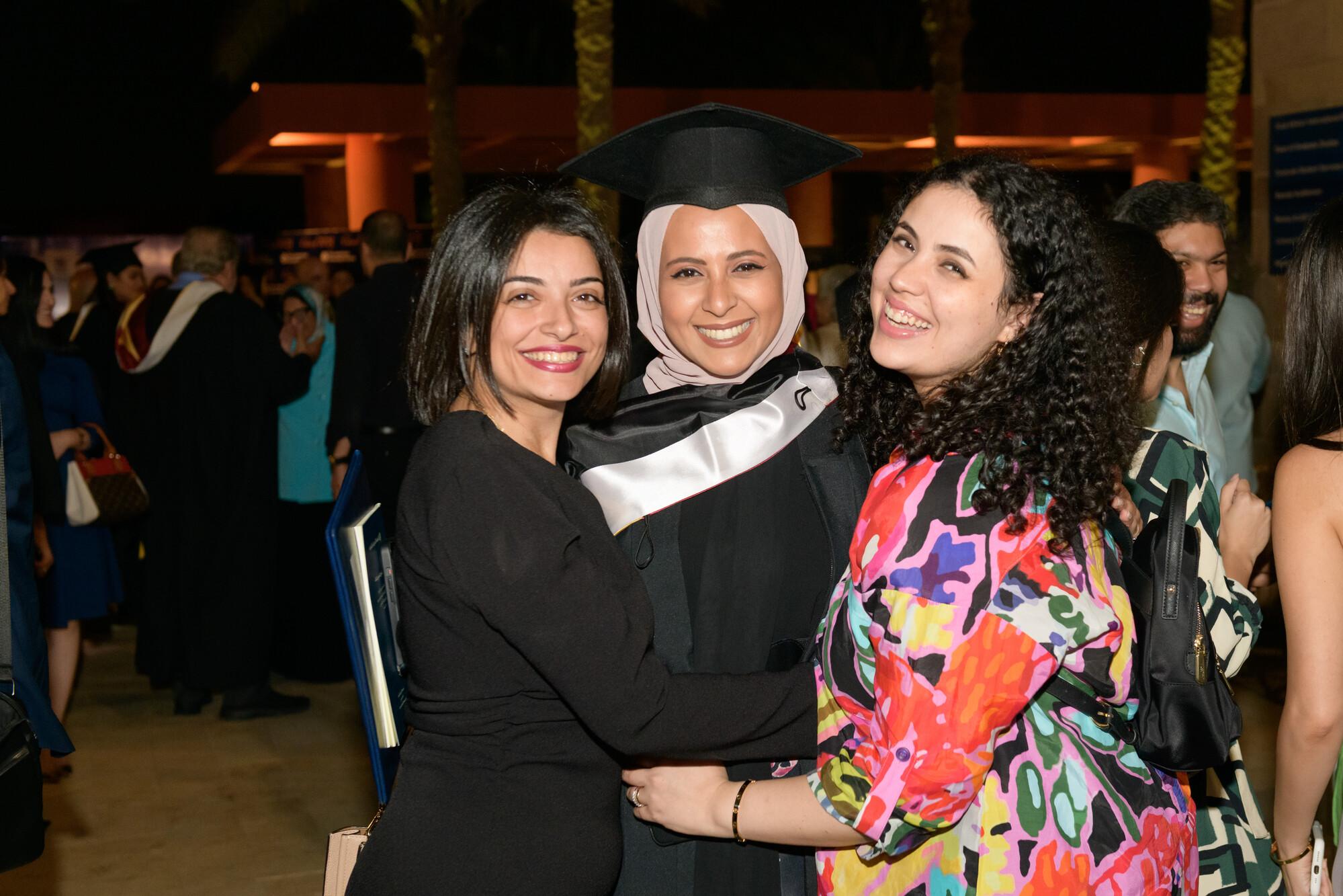 A graduating female student wearing a cap and gown with two other girls smiling