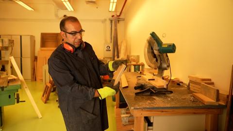 A man is working in a wood workshop. He is wearing overalls and protective glasses