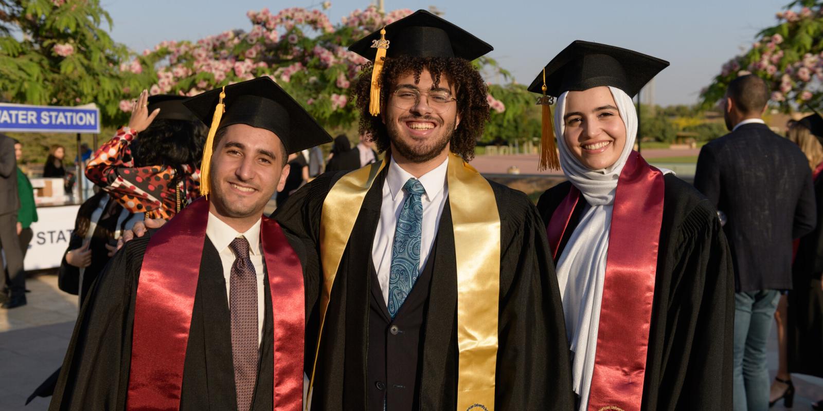 Two boys and a girl wearing a cap and gown smiling in an outdoor setting