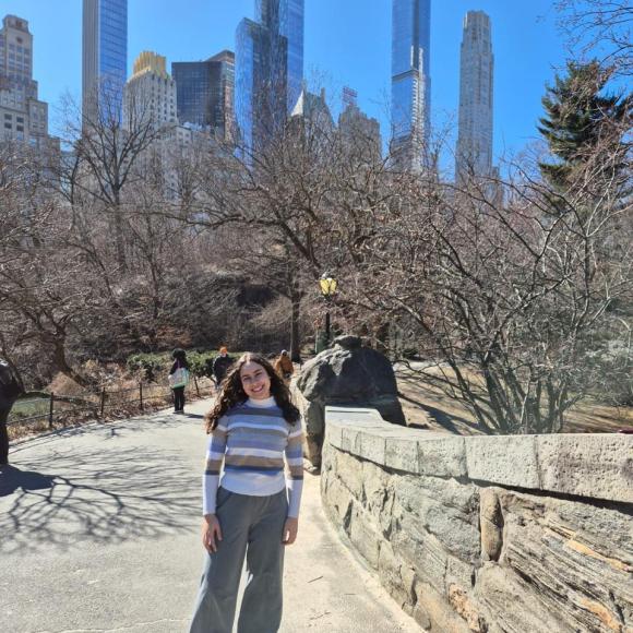 A female is standing in a park. There are sky scrapers behind her
