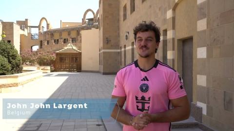 An international student is standing next to a wall and smiling. Text: John John Largent