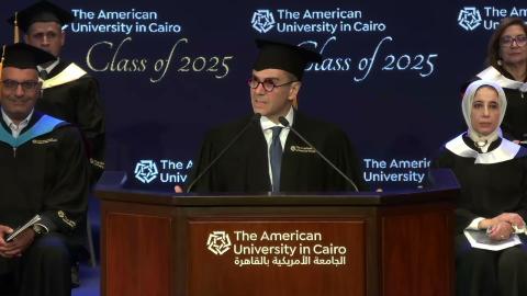 A man is wearing a cap and gown. He is standing at a podium and talking into two microphone. There are males and females sitting behind him. Text: The American University in Cairo