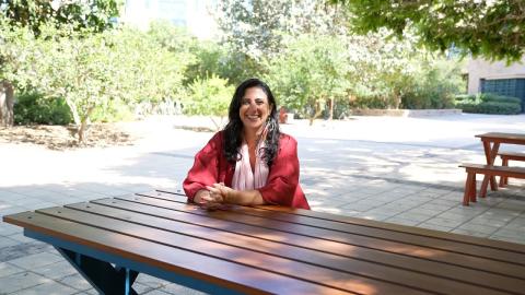Faculty Bahia Shehab is sitting at a desk outdoors and smiling
