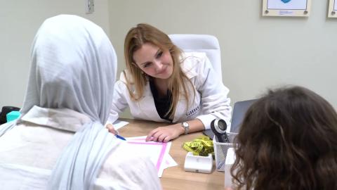 A woman is wearing a doctor's coat and sitting at a desk in front of a girl and a veiled woman