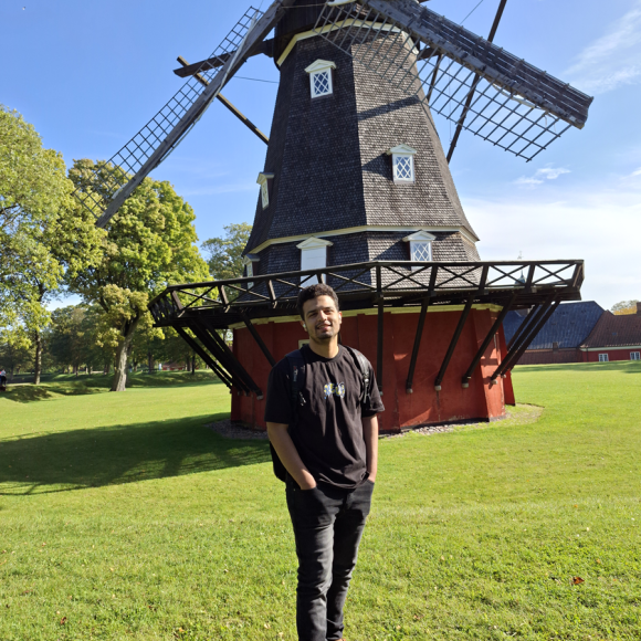 A male student is standing in front of a big windmill. He is smiling