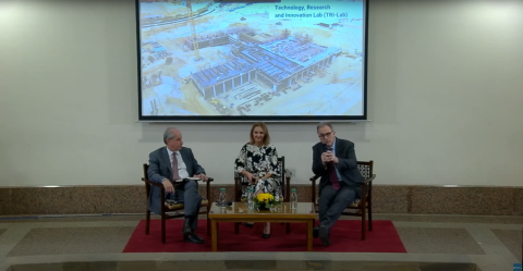 Two males and one female are sitting on a panel. There is a screen behind them displaying a construction site